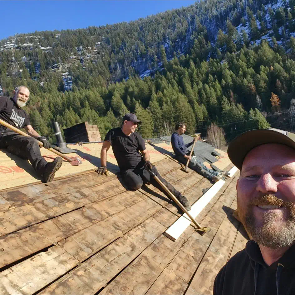 Four Trail Roofing crew members on a shingle tear-off stripped to the original plank deck, mountains behind — West Kootenays