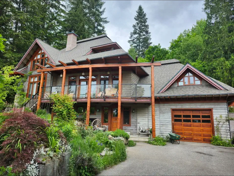 Finished grey laminate shingle roof on a multi-gable timber-frame home surrounded by gardens and evergreens — West Kootenays