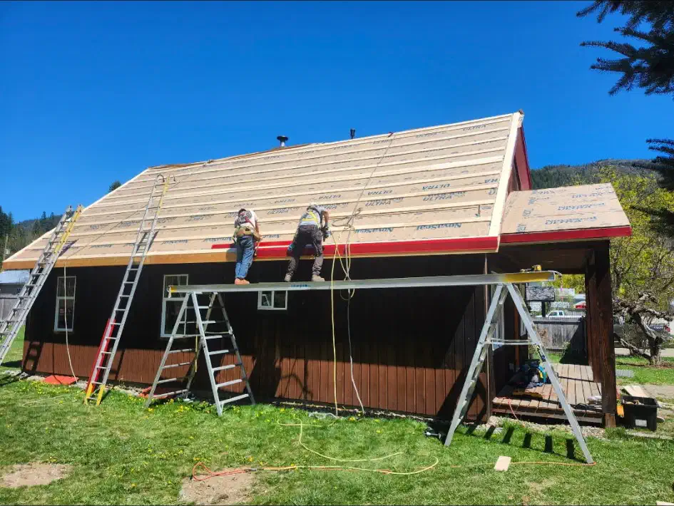 Two roofers on plank scaffolding installing new sheathing and underlayment along an eave — West Kootenays