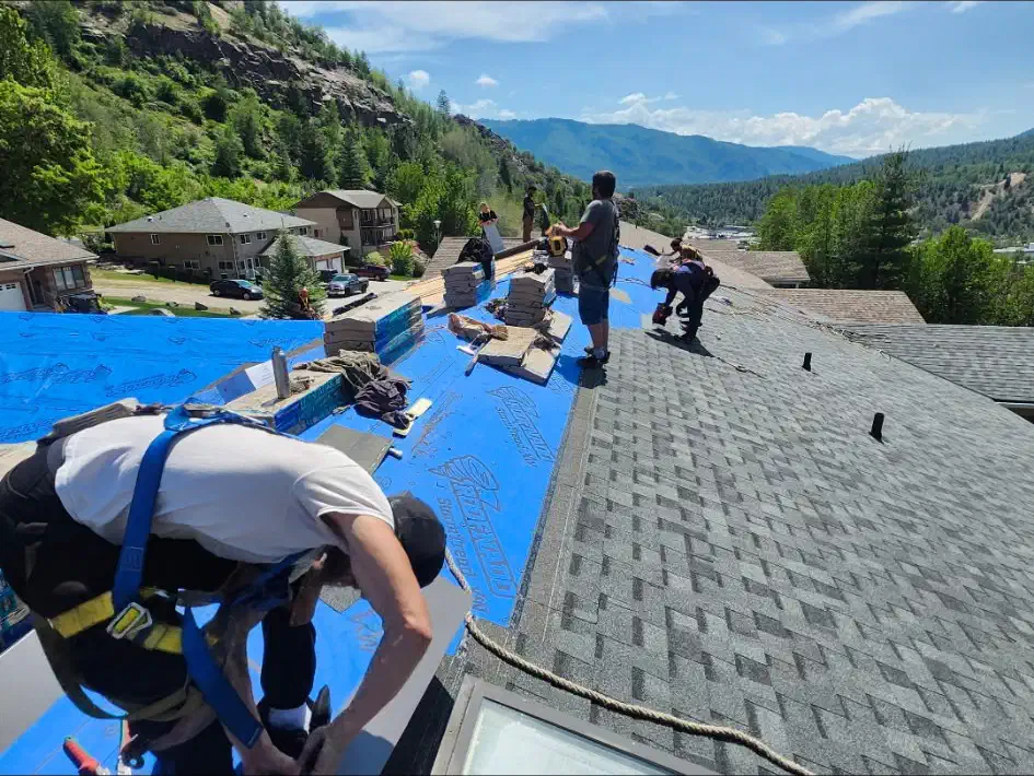 Harnessed roofing crew installs asphalt shingles over synthetic underlayment above a forested mountain valley — West Kootenays