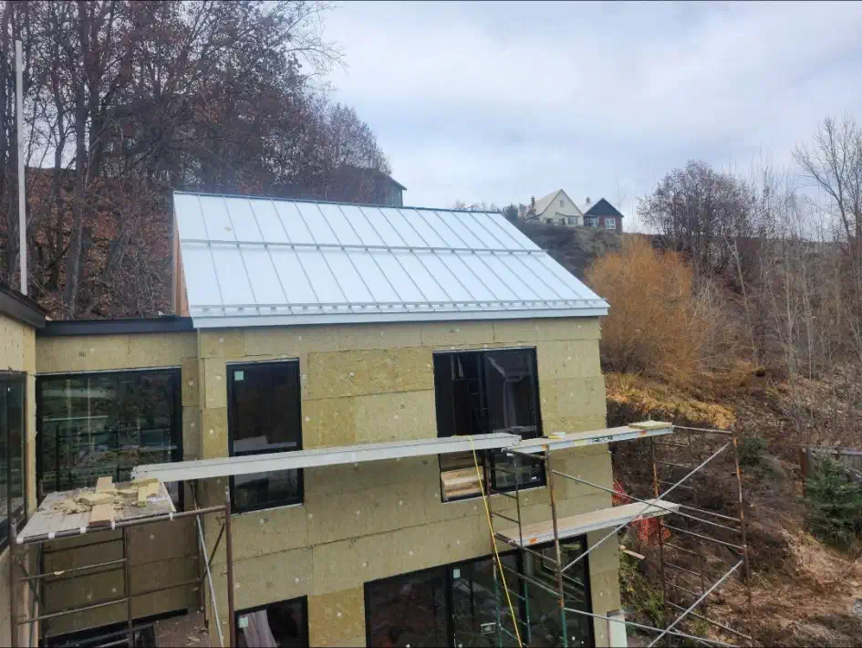Completed light-grey standing seam steel roof on a new home still wrapped in exterior insulation — West Kootenays