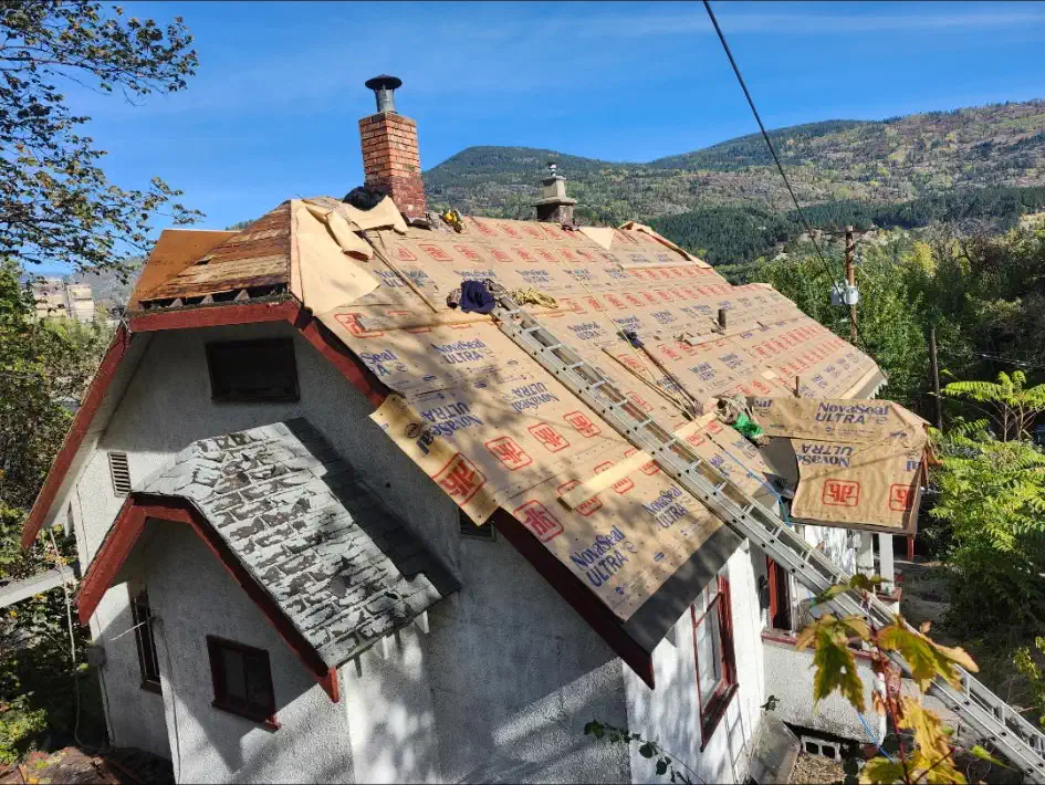 Steep roof of an older stucco home stripped and dried in with synthetic underlayment, brick chimney and valley behind — West Kootenays