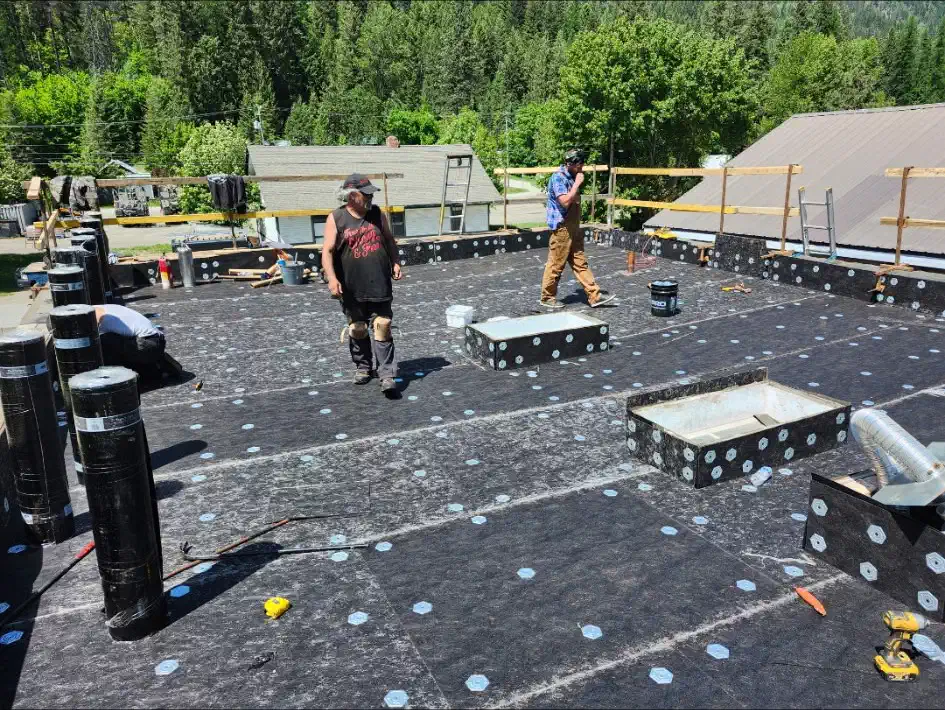 Flat roof deck covered in fastened base sheet with torch-on membrane rolls staged and guardrails installed — West Kootenays