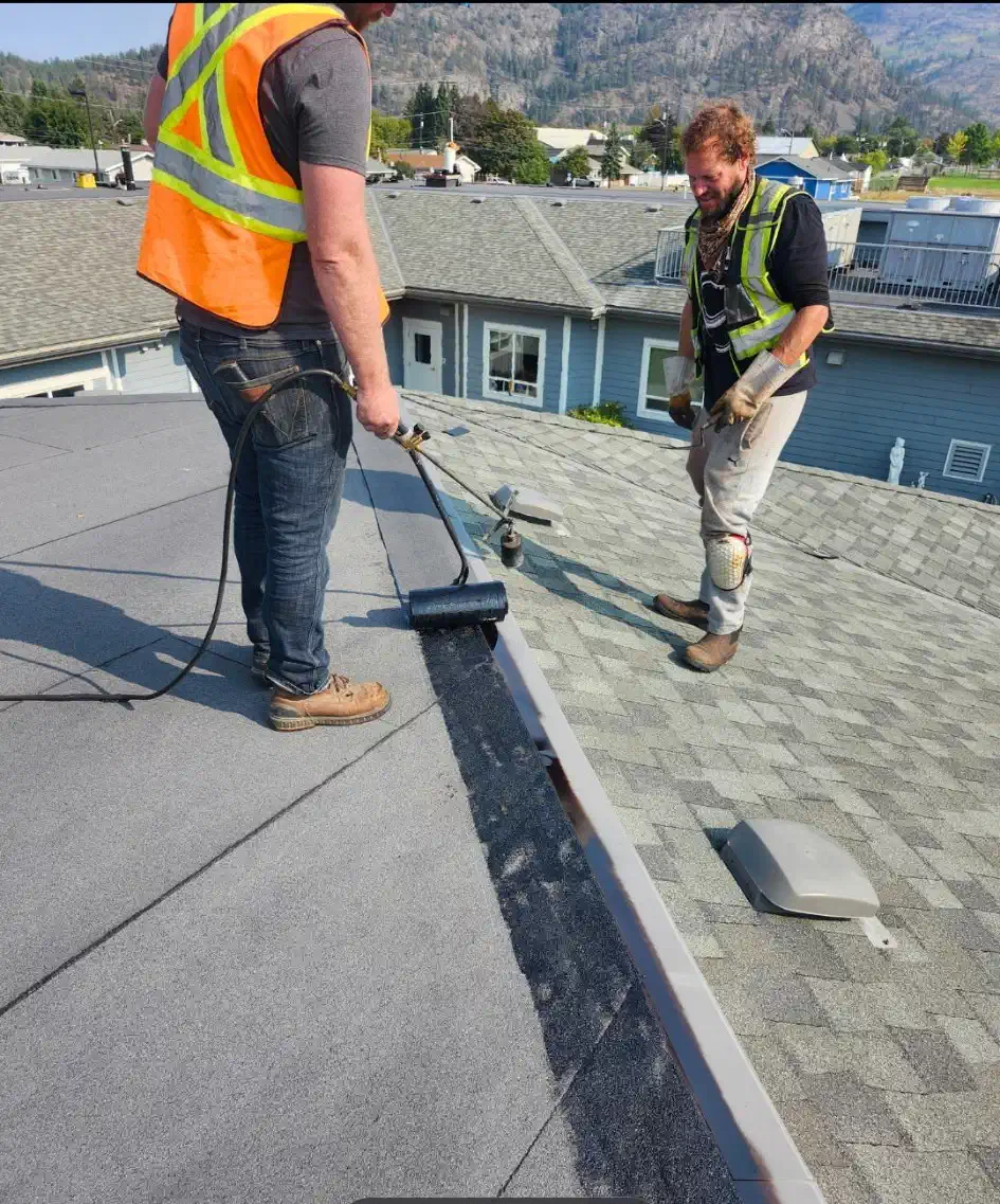 Two roofers torch-applying membrane along a metal edge where a flat roof meets a shingle slope — West Kootenays