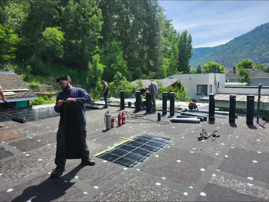 Crew staging rolls of torch-on membrane across fastened cover board on a large flat roof — West Kootenays